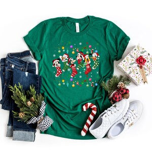 May include: A green t-shirt with a Christmas-themed design featuring Mickey Mouse, Donald Duck, Goofy, and Pluto wearing Santa hats and hanging from stockings. The design includes snowflakes, lights, and ornaments.