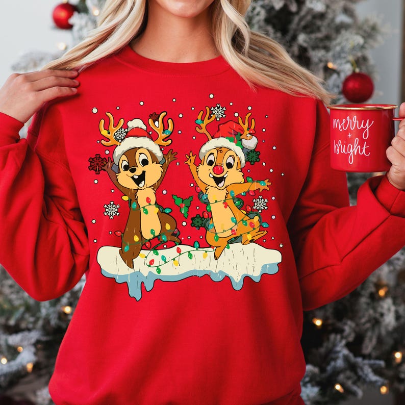 May include: Red Christmas jumper featuring a design of two chipmunks dressed as reindeer with Christmas lights. The chipmunks are wearing Santa hats and have a festive expression on their faces. The text "merry bright" is on a mug in the background.
