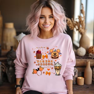 Puede incluir: Una sudadera rosa claro con un dise&ntilde;o tem&aacute;tico de Halloween. El dise&ntilde;o incluye una magdalena, una calabaza con orejas de rat&oacute;n, un caf&eacute; con leche de calabaza especiada y el texto "'Tis the Season". El dise&ntilde;o tambi&eacute;n presenta lazos naranjas y hojas de oto&ntilde;o.