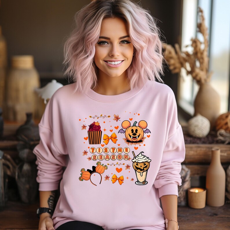 Puede incluir: Una sudadera rosa claro con un dise&ntilde;o tem&aacute;tico de Halloween. El dise&ntilde;o incluye una magdalena, una calabaza con orejas de rat&oacute;n, un caf&eacute; con leche de calabaza especiada y el texto "'Tis the Season". El dise&ntilde;o tambi&eacute;n presenta lazos naranjas y hojas de oto&ntilde;o.