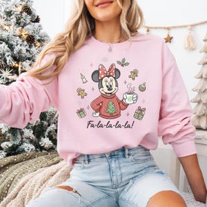 May include: A light pink sweatshirt featuring a festive Minnie Mouse design. Minnie is wearing a red sweater with a Christmas tree, holding a mug, and surrounded by holiday icons. The text "Fa-la-la-la-la!" is printed below.