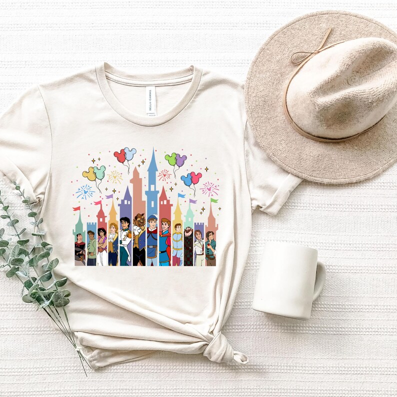 Puede incluir: Una camiseta beige con una ilustraci&oacute;n colorida de pr&iacute;ncipes de Disney de pie frente a un castillo con globos y fuegos artificiales. La camiseta tiene cuello redondo y mangas cortas.