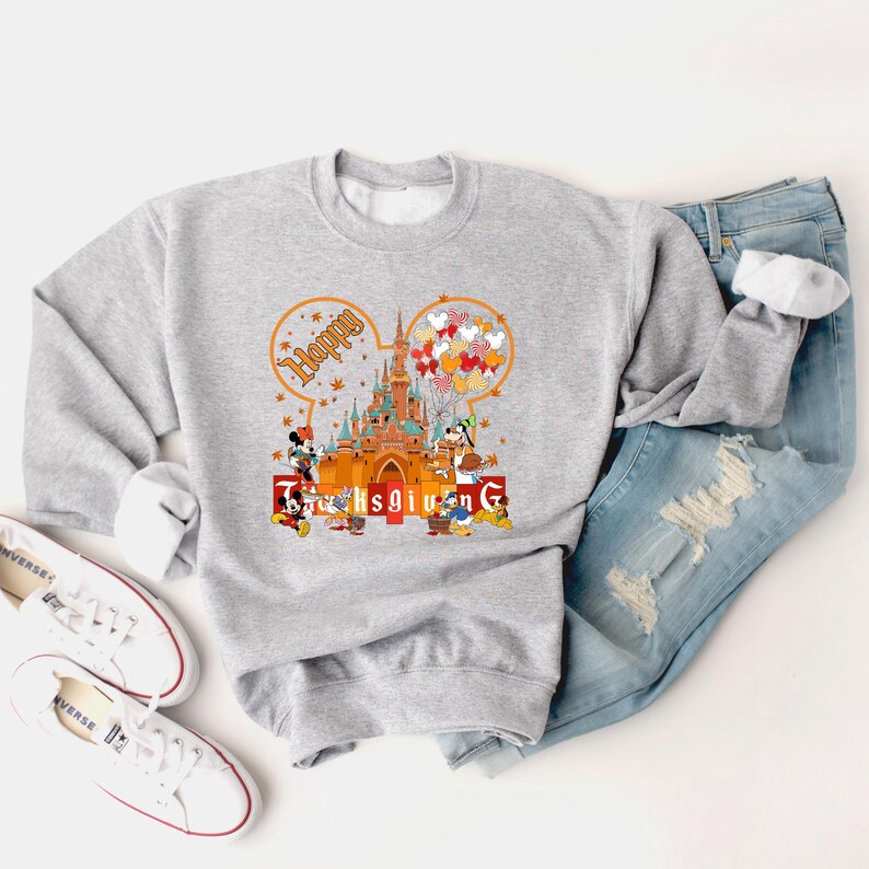 May include: A gray sweatshirt featuring a Thanksgiving design with the text "Happy Thanksgiving". The design includes a castle, cartoon characters, and fall-themed elements. The sweatshirt is paired with blue jeans and white sneakers.