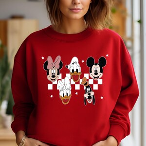 Puede incluir: Sudadera roja con un patr&oacute;n de cuadros blancos y un gr&aacute;fico de Mickey Mouse, Minnie Mouse, Donald Duck, Daisy Duck y Goofy. Los personajes sonr&iacute;en y tienen un fondo blanco.