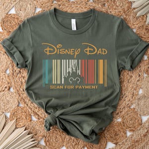 May include: A green t-shirt with a vintage-style graphic that says "Disney Dad" with a barcode underneath that has a silhouette of Cinderella's castle and Mickey Mouse. The barcode says "Scan for Payment" underneath.