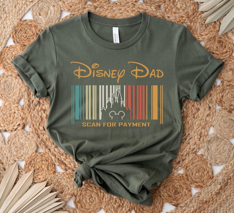 May include: A green t-shirt with a vintage-style graphic that says "Disney Dad" with a barcode underneath that has a silhouette of Cinderella's castle and Mickey Mouse. The barcode says "Scan for Payment" underneath.