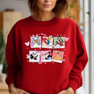K&ouml;nnte beinhalten: Roter Pullover mit einem Grafikdruck der Disney-Charaktere Mickey Mouse, Minnie Mouse, Donald Duck, Daisy Duck, Goofy und Pluto. Die Charaktere befinden sich in einzelnen Fotorahmen mit Herzen und Sternen drumherum.