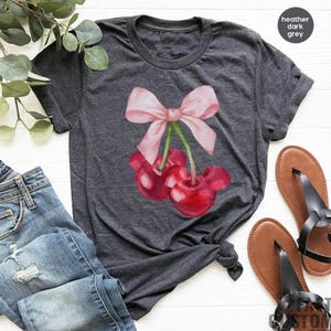 Puede incluir: Una camiseta gris oscuro jaspeada con un lazo rosa y dos cerezas rojas. Las cerezas tienen tallos con hojas y est&aacute;n atadas juntas con un lazo rosa.