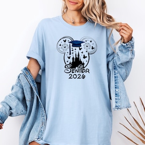 Puede incluir: Camiseta azul claro con un gr&aacute;fico negro. El gr&aacute;fico incluye un birrete, fuegos artificiales y un castillo dentro de un contorno de la cabeza de Mickey Mouse. El texto "Senior 2026" est&aacute; debajo.