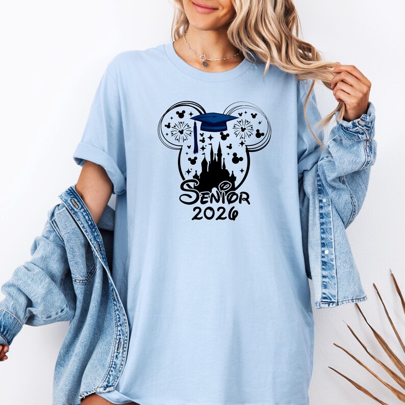 Puede incluir: Camiseta azul claro con un gr&aacute;fico negro. El gr&aacute;fico incluye un birrete, fuegos artificiales y un castillo dentro de un contorno de la cabeza de Mickey Mouse. El texto "Senior 2026" est&aacute; debajo.