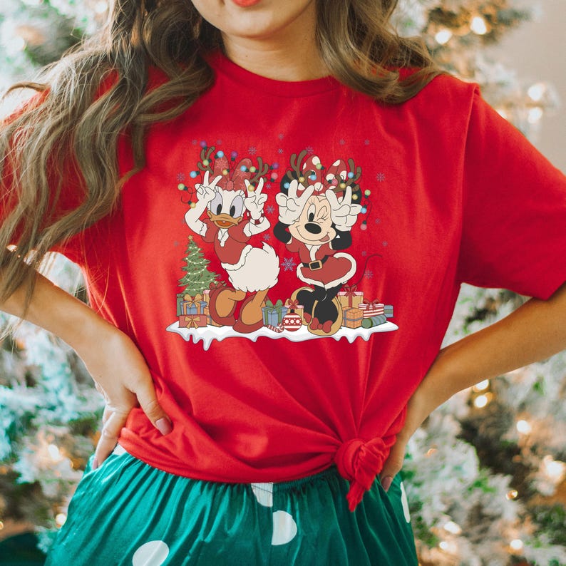 K&ouml;nnte beinhalten: Rotes T-Shirt mit einem weihnachtlichen Motiv, das Minnie Maus und Daisy Duck zeigt, die Weihnachtsm&uuml;tzen tragen und von Geschenken und einem Weihnachtsbaum umgeben sind.