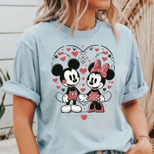 May include: Light blue t-shirt with a graphic print of Mickey Mouse and Minnie Mouse holding hands inside a heart shape. The heart is surrounded by small red hearts.