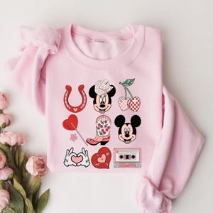 May include: Pink sweatshirt with a graphic design featuring Mickey Mouse in a cowboy hat, a horseshoe, cherries, a heart-shaped lollipop, a boot, a cassette tape, and hearts.