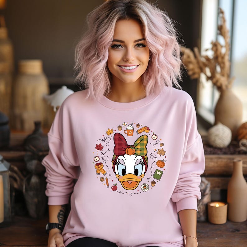Puede incluir: Sudadera rosa claro con un dise&ntilde;o de Daisy Duck con tem&aacute;tica oto&ntilde;al. El dise&ntilde;o incluye un lazo, hojas, calabazas y una taza de caf&eacute;. La sudadera es de manga larga y tiene cuello redondo.