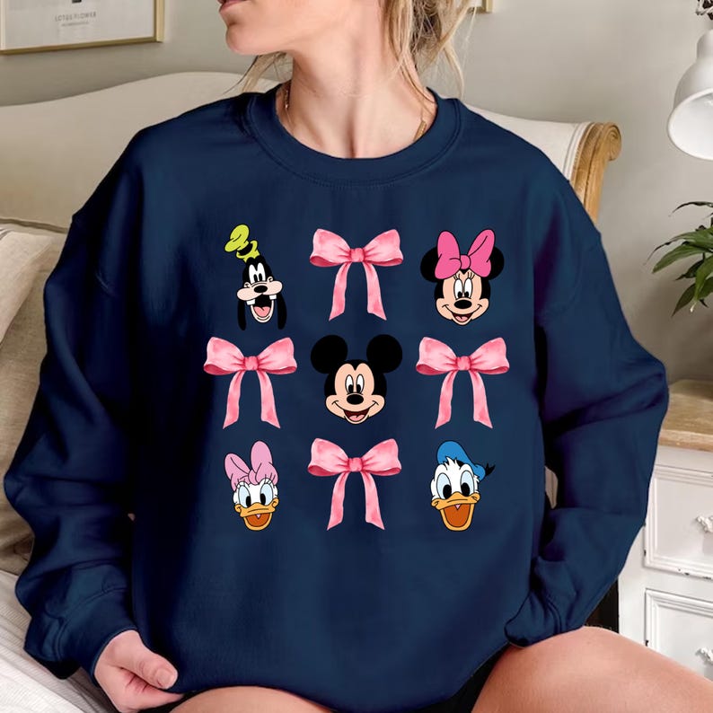 May include: A navy blue sweatshirt with a graphic print of Mickey Mouse, Minnie Mouse, Donald Duck, and Goofy. Each character is wearing a pink bow.