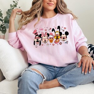 Op de afbeelding: Een roze sweatshirt met een grafische print van Mickey Mouse, Minnie Mouse, Donald Duck, Goofy en Pluto die hartvormige zonnebrillen dragen. De personages zijn omringd door harten, sterren en andere schattige details. De tekst "Love" is op het sweatshirt gedrukt.