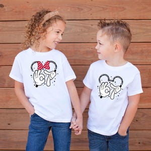 K&ouml;nnte beinhalten: Zwei Kinder tragen wei&szlig;e T-Shirts mit einem schwarz-wei&szlig;en Design im Mickey-Mouse-Stil. Ein Shirt hat eine rote Schleife mit Punkten. Das Design beinhaltet eine Handgeste und die Zahl 67. Die Kinder halten sich an den H&auml;nden.