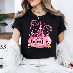 May include: Black t-shirt with a pink and white graphic of Mickey Mouse and friends celebrating Valentine's Day at a pink castle with the word "Valentine" in pink letters.