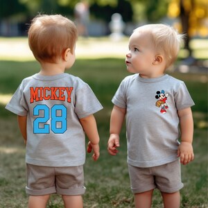 May include: Two toddlers wearing grey t-shirts. One shirt has the text "MICKEY 28" on the back. The other shirt has a cartoon image of Mickey Mouse playing baseball.