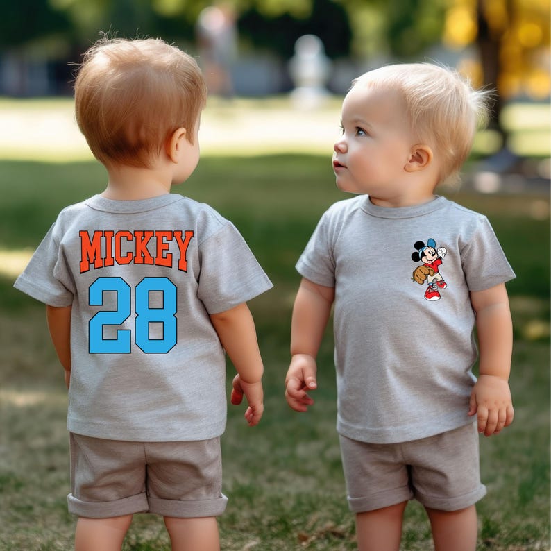 May include: Two toddlers wearing grey t-shirts. One shirt has the text "MICKEY 28" on the back. The other shirt has a cartoon image of Mickey Mouse playing baseball.