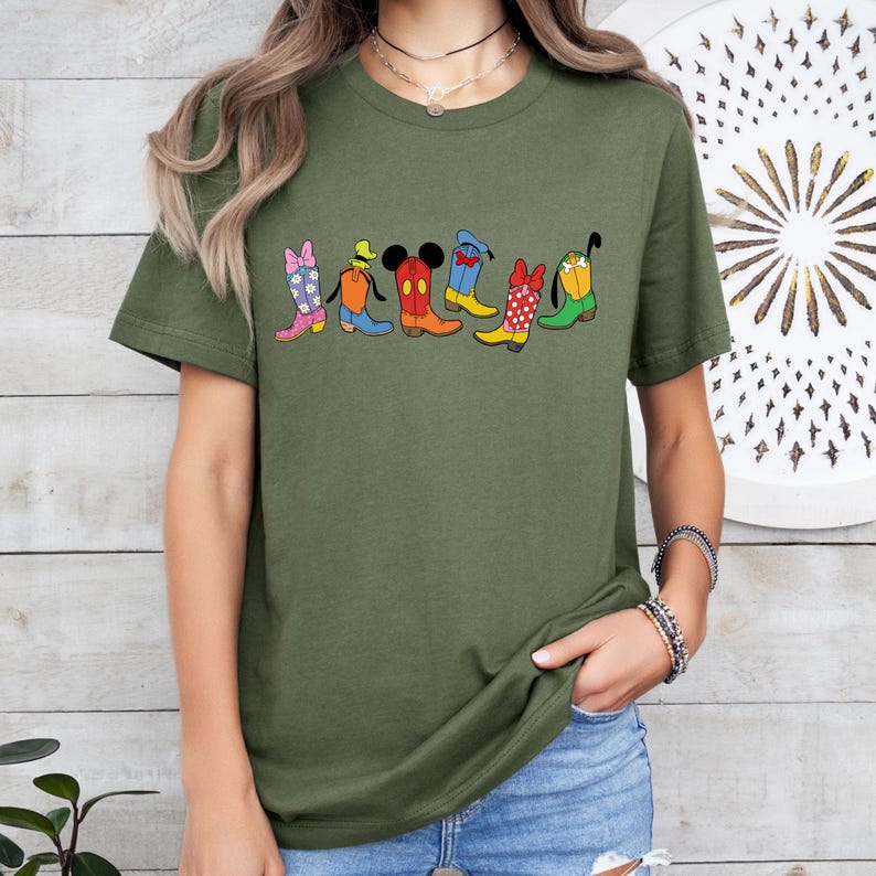 May include: Olive green t-shirt featuring a row of cartoon cowboy boots in various colors and patterns. The boots have character-inspired designs, including Mickey Mouse and Minnie Mouse, with bows, polka dots, and other details.