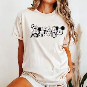 K&ouml;nnte beinhalten: Ein cremefarbenes T-Shirt mit einer schwarz-wei&szlig;en Grafik von Mickey Mouse, Minnie Mouse, Donald Duck, Goofy und Pluto. Die Figuren stehen in einer Reihe und ihre Gesichter sind alle sichtbar.