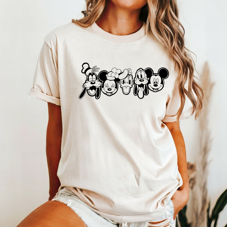 K&ouml;nnte beinhalten: Ein cremefarbenes T-Shirt mit einer schwarz-wei&szlig;en Grafik von Mickey Mouse, Minnie Mouse, Donald Duck, Goofy und Pluto. Die Figuren stehen in einer Reihe und ihre Gesichter sind alle sichtbar.