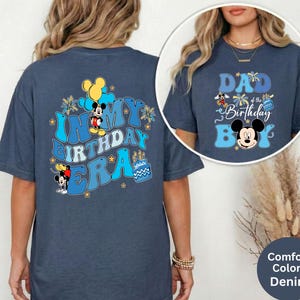 Op de afbeelding: Denimblauwe T-shirts met Mickey Mouse-afbeeldingen. E&eacute;n shirt toont "IN MY BIRTHDAY ERA" met Mickey Mouse en ballonnen. De andere zegt "DAD of the Birthday Boy" met Mickey Mouse. De shirts maken deel uit van de Comfort Colors-collectie.
