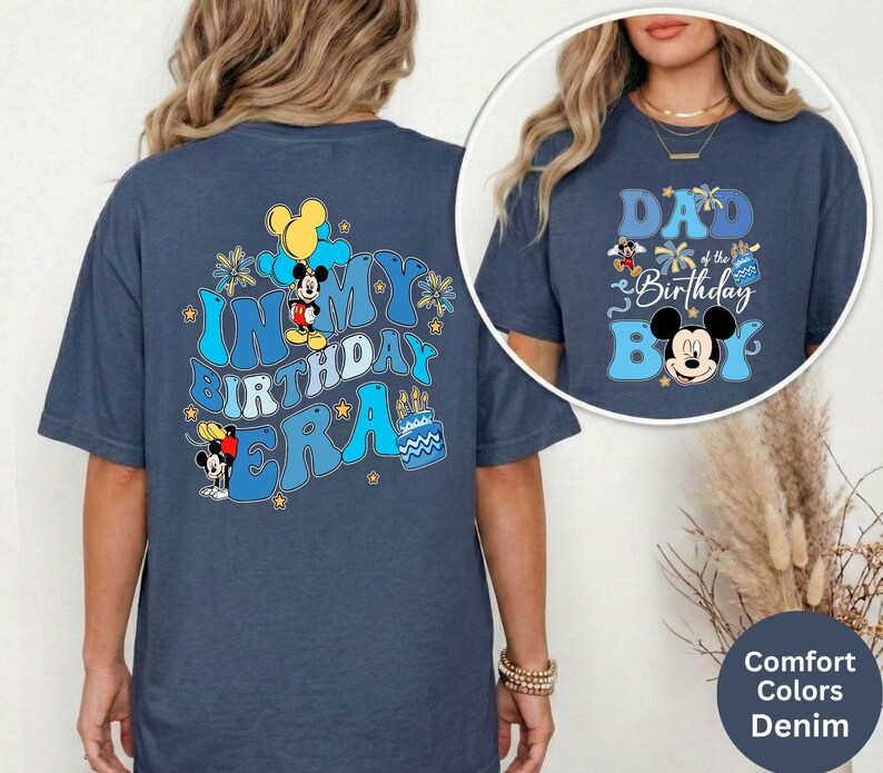 Op de afbeelding: Denimblauwe T-shirts met Mickey Mouse-afbeeldingen. E&eacute;n shirt toont "IN MY BIRTHDAY ERA" met Mickey Mouse en ballonnen. De andere zegt "DAD of the Birthday Boy" met Mickey Mouse. De shirts maken deel uit van de Comfort Colors-collectie.