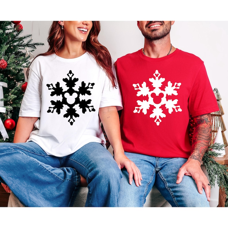 May include: White and red t-shirts featuring a snowflake design with a Mickey Mouse head in the center. The black snowflake is on the white shirt, and the white snowflake is on the red shirt. Both shirts are paired with blue jeans.