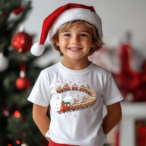 May include: A child wearing a white t-shirt with a Christmas train graphic and a red Santa hat. The train features gingerbread people and animals in each car, with snowflakes around the design. The background includes a Christmas tree and ornaments.