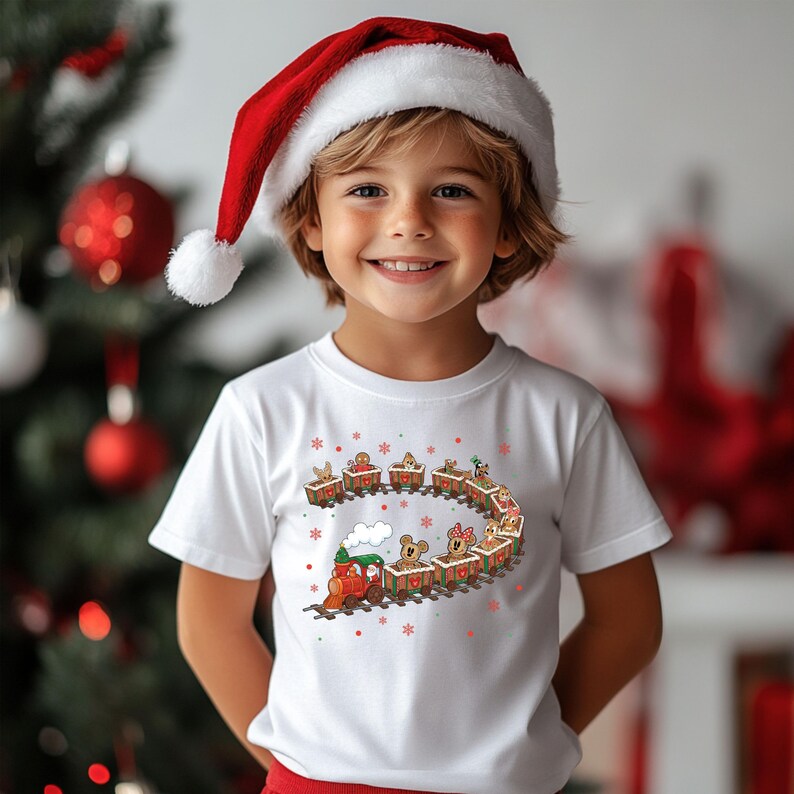 May include: A child wearing a white t-shirt with a Christmas train graphic and a red Santa hat. The train features gingerbread people and animals in each car, with snowflakes around the design. The background includes a Christmas tree and ornaments.
