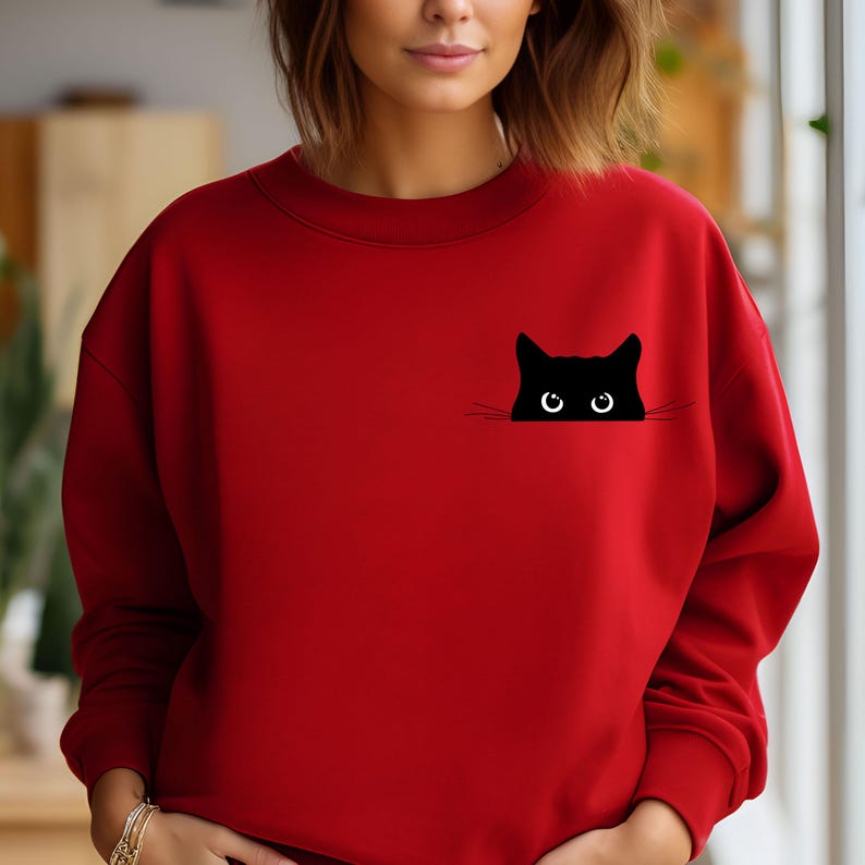 Pu&ograve; includere: Felpa rossa con un gatto nero che spunta dalla tasca. Il gatto ha grandi occhi rotondi.
