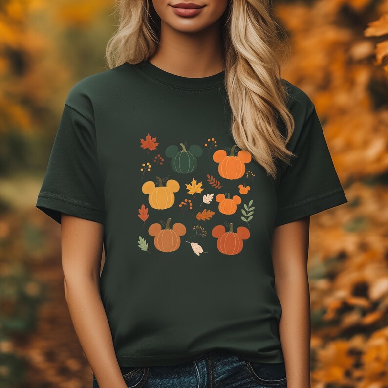 May include: Dark green t-shirt featuring a fall-themed design. The design includes cartoon pumpkins in orange, yellow, and green, with Mickey Mouse ears, surrounded by autumn leaves. A seasonal graphic tee.