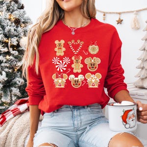 May include: Red Christmas sweater with a gingerbread Mickey Mouse and Minnie Mouse design. The sweater features a variety of gingerbread cookies, including candy canes, a gingerbread house, and a gingerbread ornament.