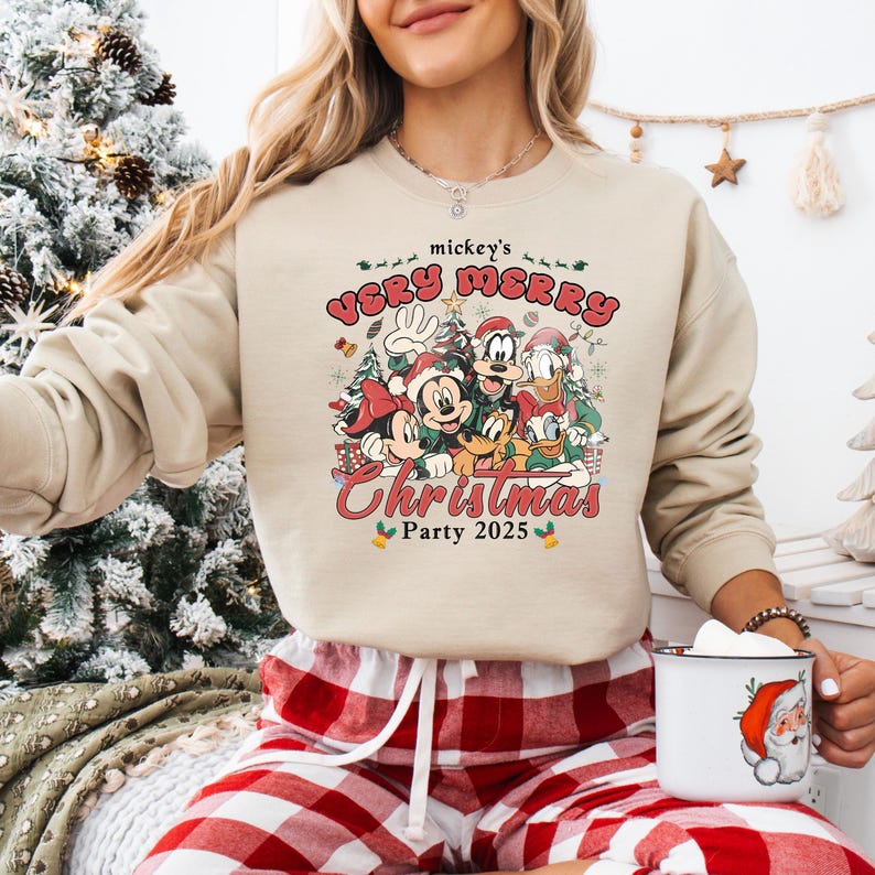 Puede incluir: Sudadera beige con un gr&aacute;fico colorido de Mickey Mouse, Minnie Mouse, Donald, Goofy y Pluto celebrando la Navidad. El texto "mickey's VERY MERRY Christmas Party 2025" est&aacute; impreso encima y debajo de los personajes.