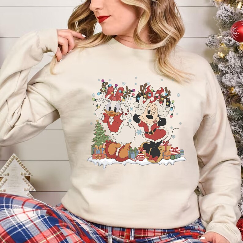 K&ouml;nnte beinhalten: Ein cremefarbener Pullover mit einem weihnachtlichen Design, das Minnie Mouse und Daisy Duck als Weihnachtsmann zeigt. Sie tragen rot-wei&szlig;e Weihnachtsmannkost&uuml;me mit Rentiergeweihen und Weihnachtslichtern. Das Design ist von Geschenken und einem kleinen Weihnachtsbaum umgeben.