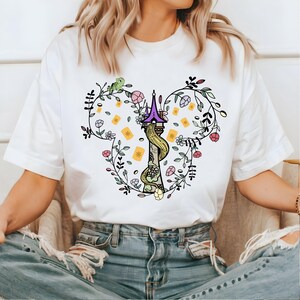 May include: White t-shirt with a floral design featuring a purple tower with a long blonde braid, surrounded by yellow lanterns and green leaves.