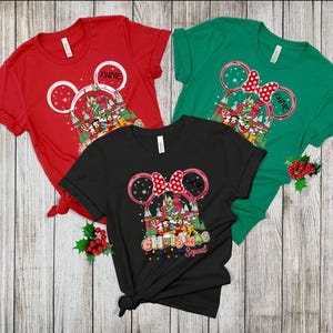 Puede incluir: Tres camisetas de manga corta en rojo, verde y negro. Cada camiseta presenta un dise&ntilde;o de orejas de Mickey Mouse con tem&aacute;tica navide&ntilde;a, incluyendo un castillo, personajes y el texto "Christmas Squad". La camiseta roja tiene espacio para un nombre.