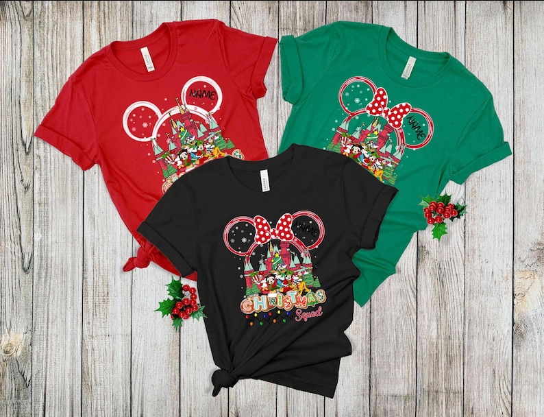 Puede incluir: Tres camisetas de manga corta en rojo, verde y negro. Cada camiseta presenta un dise&ntilde;o de orejas de Mickey Mouse con tem&aacute;tica navide&ntilde;a, incluyendo un castillo, personajes y el texto "Christmas Squad". La camiseta roja tiene espacio para un nombre.
