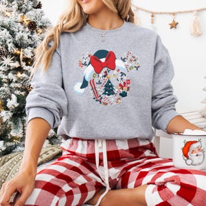 May include: A grey sweatshirt with a Christmas-themed design, including a Santa hat, red bow, and Christmas tree. The design incorporates festive elements. The person is wearing red and white plaid pyjama bottoms and holding a mug with a Santa Claus graphic.