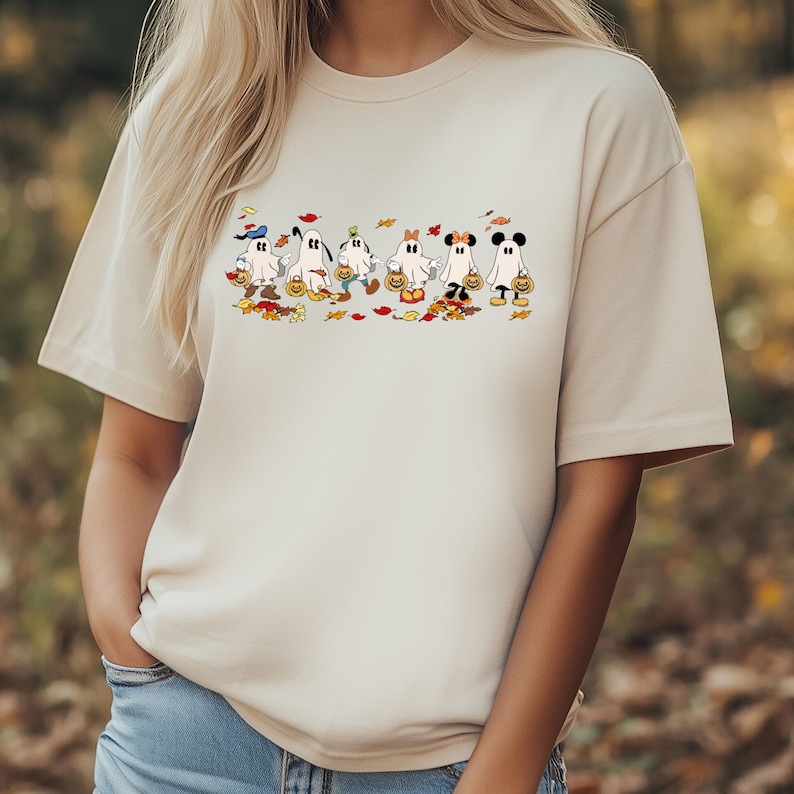 May include: Beige t-shirt featuring a Halloween-themed graphic. The graphic depicts cartoon ghosts dressed as popular characters, each holding a pumpkin-shaped candy bucket. Autumn leaves are scattered around the ghosts.