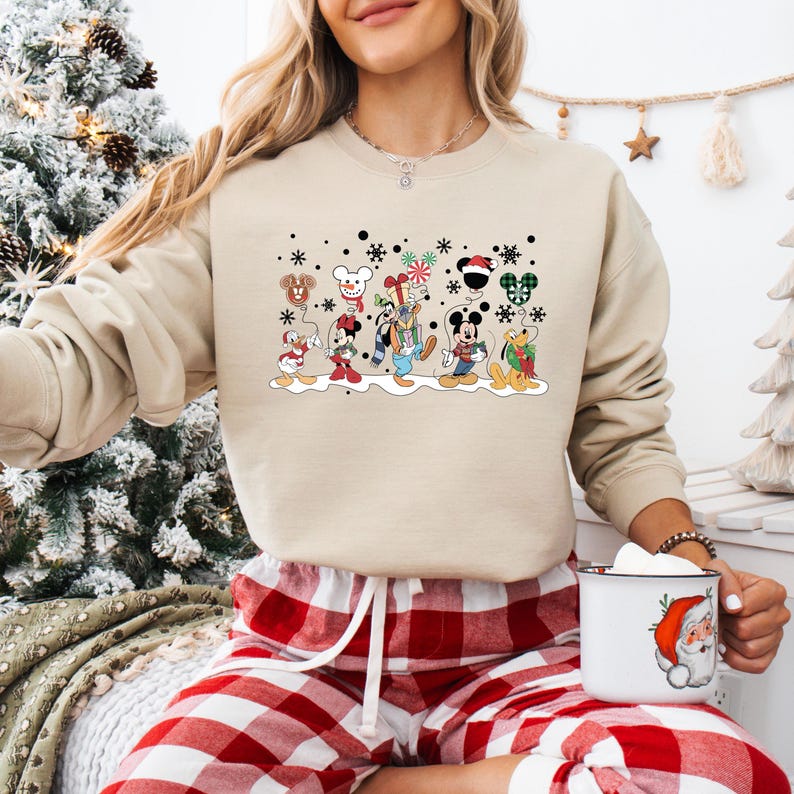 May include: A beige sweatshirt with a festive design featuring Mickey Mouse and his friends celebrating Christmas. The characters are holding balloons and gifts, and there are snowflakes in the background.