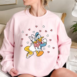 May include: Pink sweatshirt with a graphic print of Donald Duck and Daisy Duck holding hands surrounded by pink hearts.
