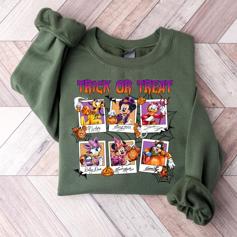 May include: Olive green sweatshirt featuring a Halloween-themed graphic. The design includes the text "Trick or Treat" in purple and orange, along with images of Disney characters dressed in Halloween costumes. The characters include Mickey Mouse, Donald Duck, and Goofy.