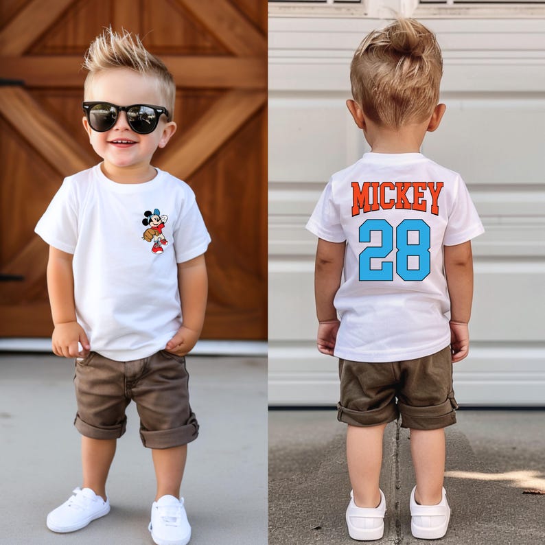 May include: A white t-shirt with a Mickey Mouse graphic on the front and the text "Mickey 28" on the back. The shirt is being worn by a young boy with brown hair and sunglasses. He is also wearing khaki shorts.
