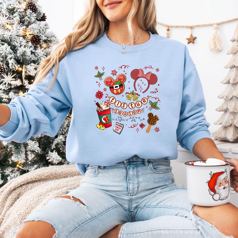 May include: Light blue sweatshirt with a festive graphic featuring Mickey Mouse, a hot chocolate, and the phrase "'Tis the Season." The design includes holly, bells, and a Mickey Mouse ear headband. The person is holding a Santa mug.