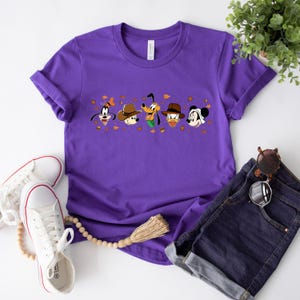 May include: A purple t-shirt featuring cartoon characters Goofy, Mickey Mouse, Donald Duck, and Pluto wearing hats. The graphic includes autumn leaves. The shirt is paired with denim shorts, sunglasses, and white sneakers.