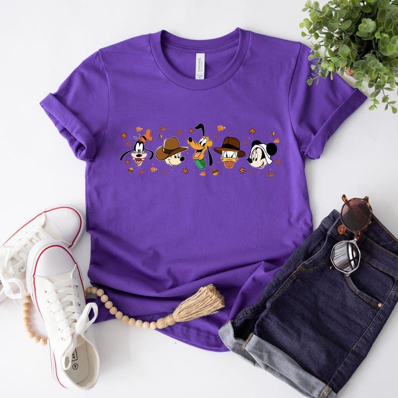May include: A purple t-shirt featuring cartoon characters Goofy, Mickey Mouse, Donald Duck, and Pluto wearing hats. The graphic includes autumn leaves. The shirt is paired with denim shorts, sunglasses, and white sneakers.