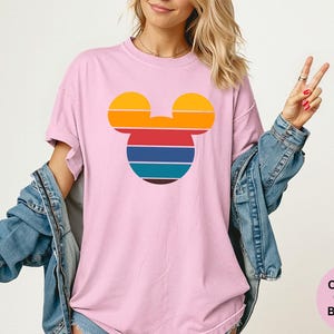 K&ouml;nnte beinhalten: Hellrosa T-Shirt mit einem Retro-Mickey-Mouse-Design. Das Design ist eine Silhouette von Mickeys Kopf mit horizontalen Streifen in Gelb, Orange, Rot, Blau und T&uuml;rkis. Das Shirt ist ein Comfort Colors Blossom.